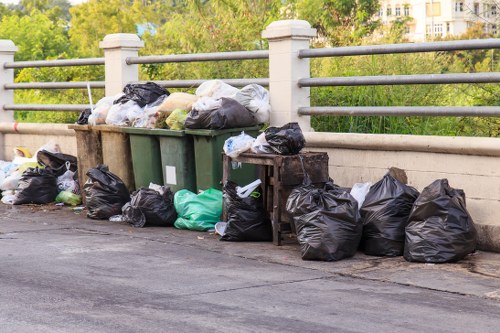 A row of black, green, and white rubbish bags filled with household waste are placed along a concrete pavement, with some bags leaning against a green wheelie bin that is positioned underneath a metal railing. Additional rubbish bags are stacked on the ground next to the bin, which appears to be near a bridge or road barrier, with a background of leafy trees and distant buildings suggesting an urban setting in Highgate or nearby London postcode areas. The bags vary in size and material, with some tightly knotted and others loosely gathered, indicating a collection ready for waste removal. The scene captures the outdoor environment on a clear day, with natural light illuminating the rubbish and the surrounding area subtly indicating a neighbourhood setting consistent with garden clearance and general rubbish collection services in the local area. This image visually supports professional rubbish removal services offered in Highgate by Garden Clearance Highgate.