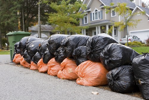 A long row of black and orange heavy-duty rubbish bags filled with waste material, stacked and lined up along the edge of a residential street. The bags are placed on the pavement in front of a suburban property with a driveway, grass lawn, and trees with green foliage. The background features a modern, multi-storey house with light grey and white exterior finishes, large windows, and a pitched roof. The scene suggests a garden or household rubbish clearance, typical of services provided by Garden Clearance Highgate in the local area, possibly in or near the N6 postcode. The sky is overcast, and the street appears clean and well-maintained, highlighting the typical scene of waste awaiting collection in a suburban setting, indicating a scheduled rubbish removal service.