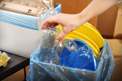 A person's hand holding a clear plastic water bottle above a blue recycling bag filled with various plastic items, including bright yellow and blue plastic buckets, situated indoors on a dark surface with a white cardboard box and wooden furniture visible in the background. The contents are stacked and partially compressed, indicating they are ready for disposal or recycling, with the scene reflecting an environment where rubbish removal services, such as those provided by Garden Clearance Highgate, might be engaged for waste collection and disposal within a residential or commercial setting in Highgate or nearby areas of London. The plastic objects have smooth textures and vibrant colours, contrasting with the neutral tones of the surroundings, emphasizing the focus on waste materials and their sorting process.
