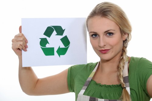 A young woman with blonde hair styled in a side braid is holding a white sign with a large green recycling symbol composed of three arrows forming a triangle. She is wearing a green short-sleeved top and a grey and white striped apron, standing against a plain white background. The image appears to relate to waste management or recycling awareness, consistent with rubbish removal themes. The woman's facial expression is neutral with a slight smile, and she is positioned slightly to the right of the frame, prominently displaying the recycling symbol. This visual could be associated with environmentally conscious rubbish clearance services in Highgate, emphasizing recycling and eco-friendly disposal practices. The clean and simple composition highlights the message of waste separation and responsible disposal, relevant to a rubbish removal service website such as garden clearance highgate.co.uk.